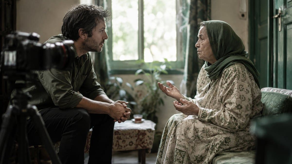 A filmmaker leaning forward listening to a woman seated across from him, camera on a tripod beside them