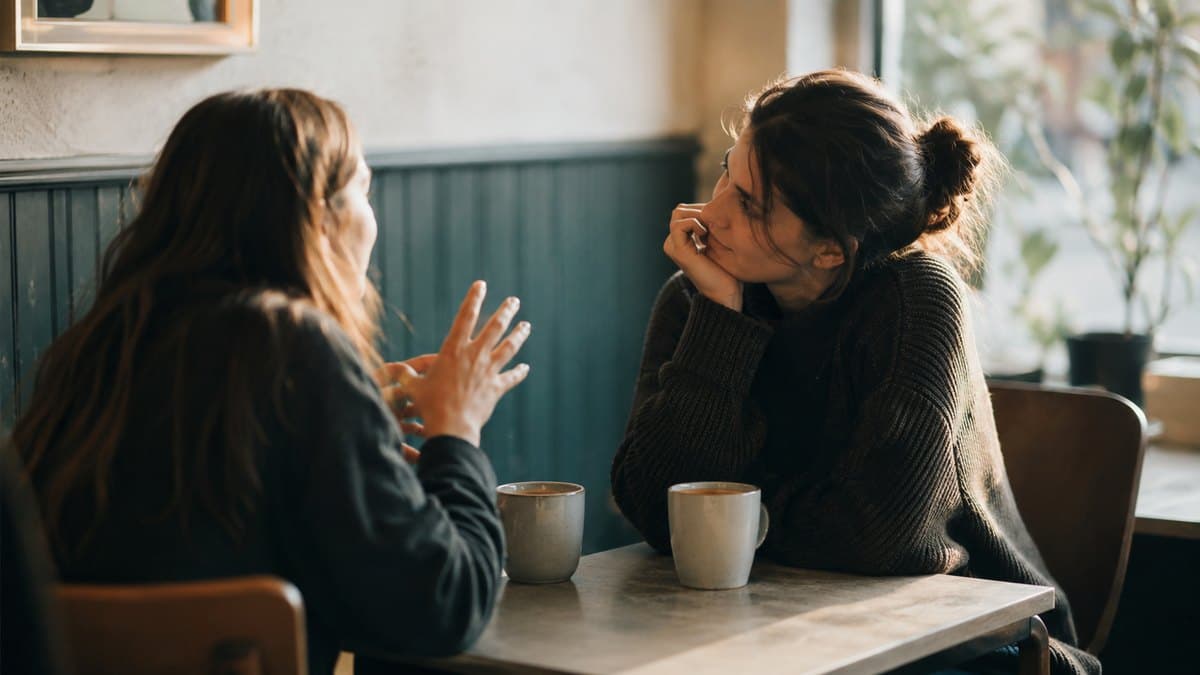 Two people talking across a table in a coffee shop, one leaning in and listening
